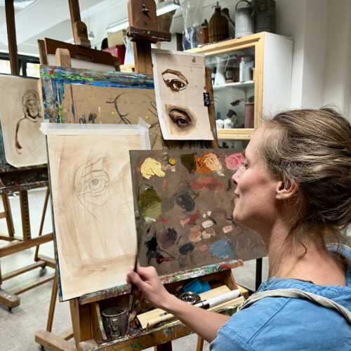 Vrouw in een kunstatelier bekijkt een schets van een oog op een ezel, met een kleurenpalet in de hand