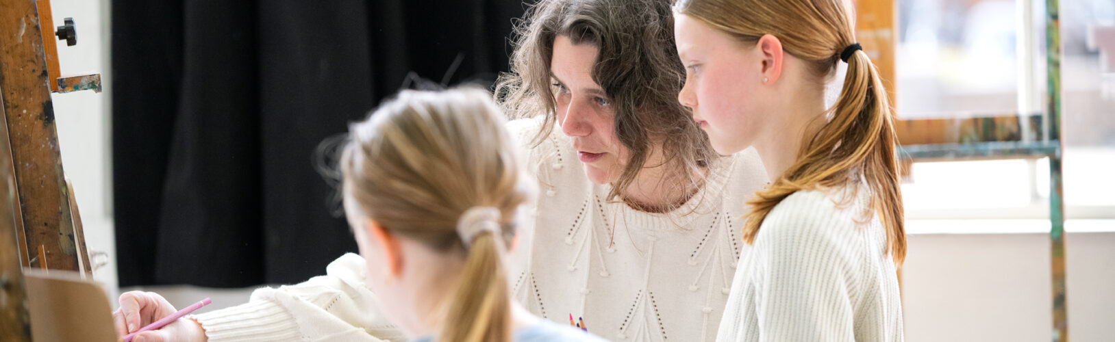 Een docent wijst met een potlood naar een schildersezel en geeft instructie aan twee meisjes in een kunstatelier