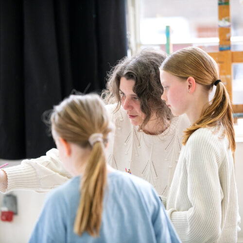 Een docent wijst met een potlood naar een schildersezel en geeft instructie aan twee meisjes in een kunstatelier