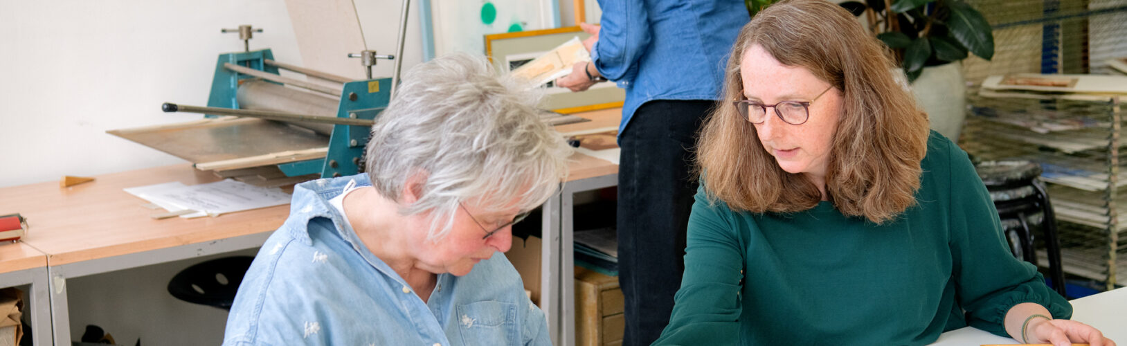 Twee vrouwen bekijken grafische kunstwerken aan een werktafel in een atelier, terwijl een derde vrouw op de achtergrond werkt bij een drukpers