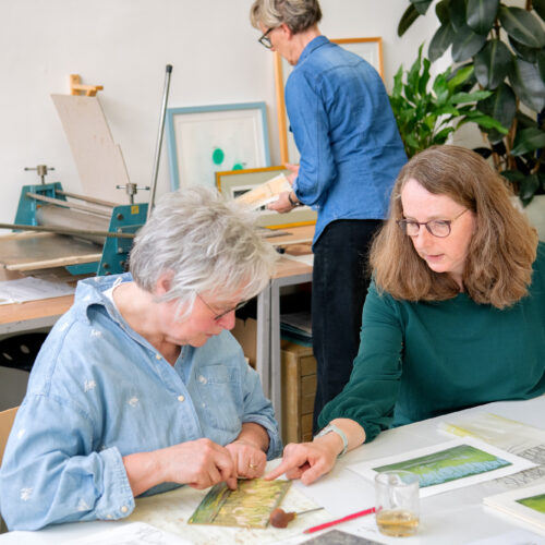 Twee vrouwen bekijken grafische kunstwerken aan een werktafel in een atelier, terwijl een derde vrouw op de achtergrond werkt bij een drukpers