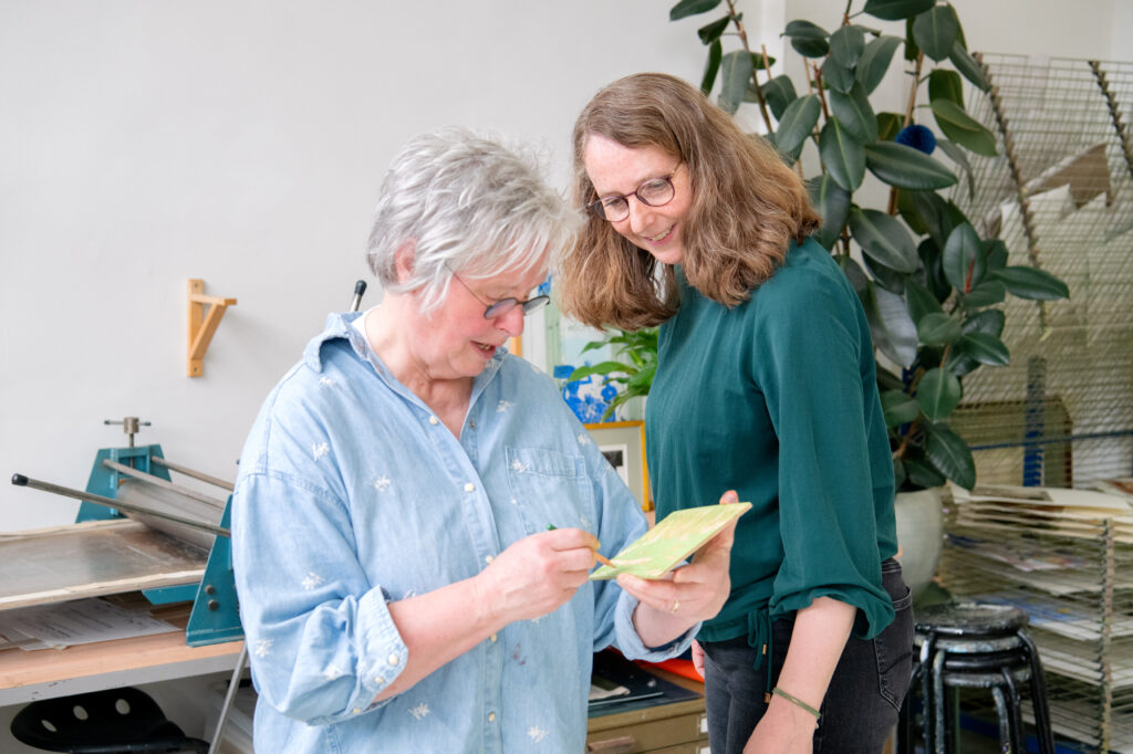 Twee vrouwen bekijken samen een klein groen kunstwerkje in een atelier met een drukpers op de achtergrond