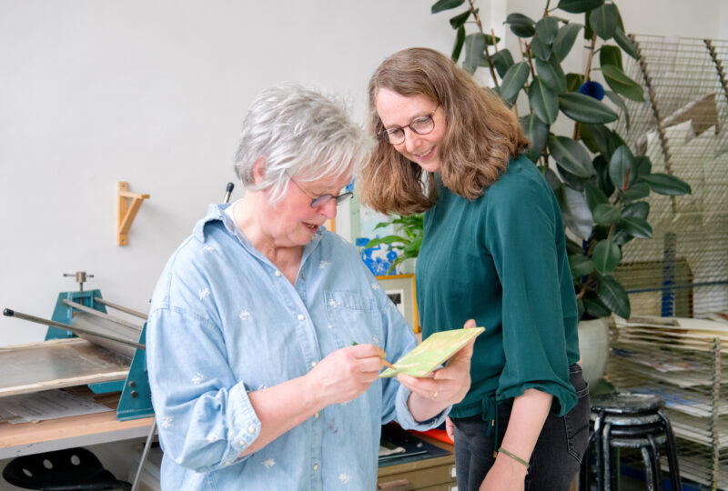 Twee vrouwen bekijken samen een klein groen kunstwerkje in een atelier met een drukpers op de achtergrond