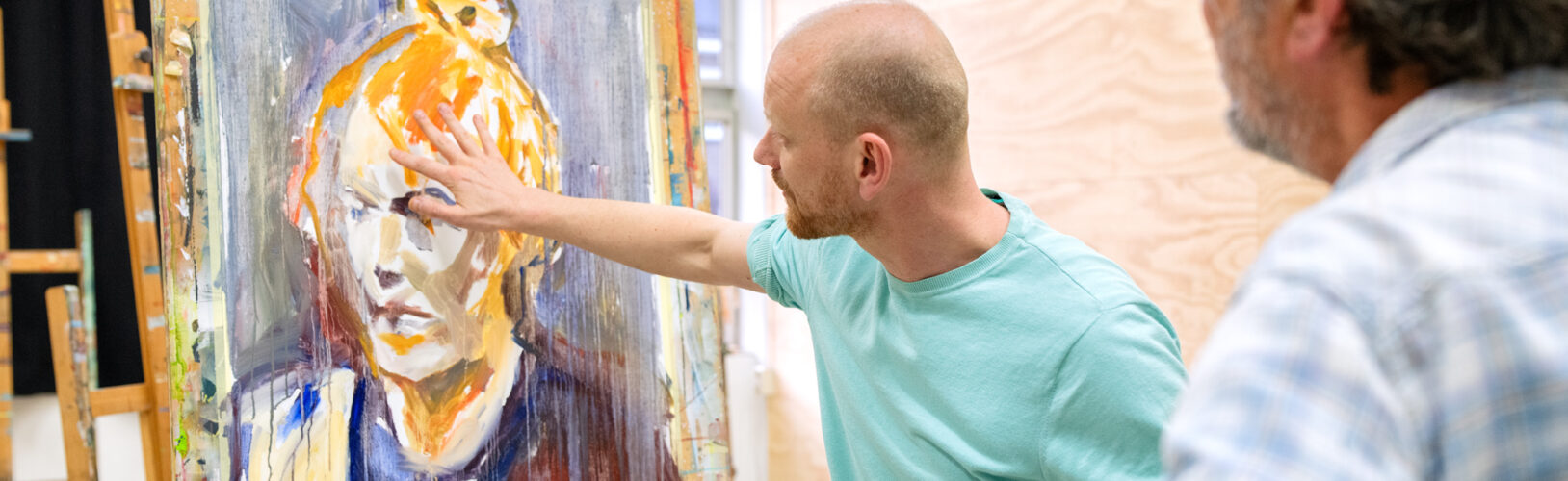 Man wijst naar een expressief geschilderd portret op een ezel in een kunstatelier, terwijl een andere persoon toekijkt