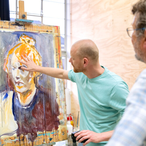 Man wijst naar een expressief geschilderd portret op een ezel in een kunstatelier, terwijl een andere persoon toekijkt