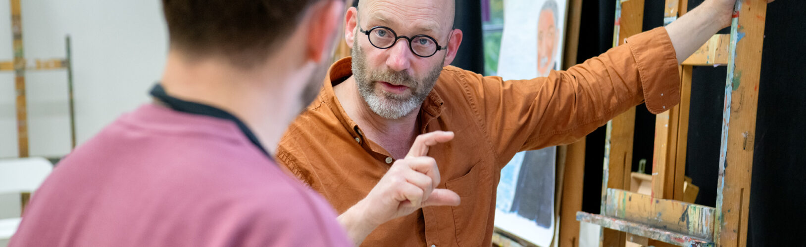 Docent geeft feedback aan student bij een schildersezel met een portretstuk in een kunstatelier