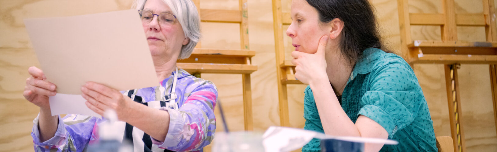 Twee vrouwen bekijken samen een vel papier aan een werktafel in een kunstatelier met schildersezels op de achtergrond