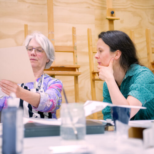 Twee vrouwen bekijken samen een vel papier aan een werktafel in een kunstatelier met schildersezels op de achtergrond